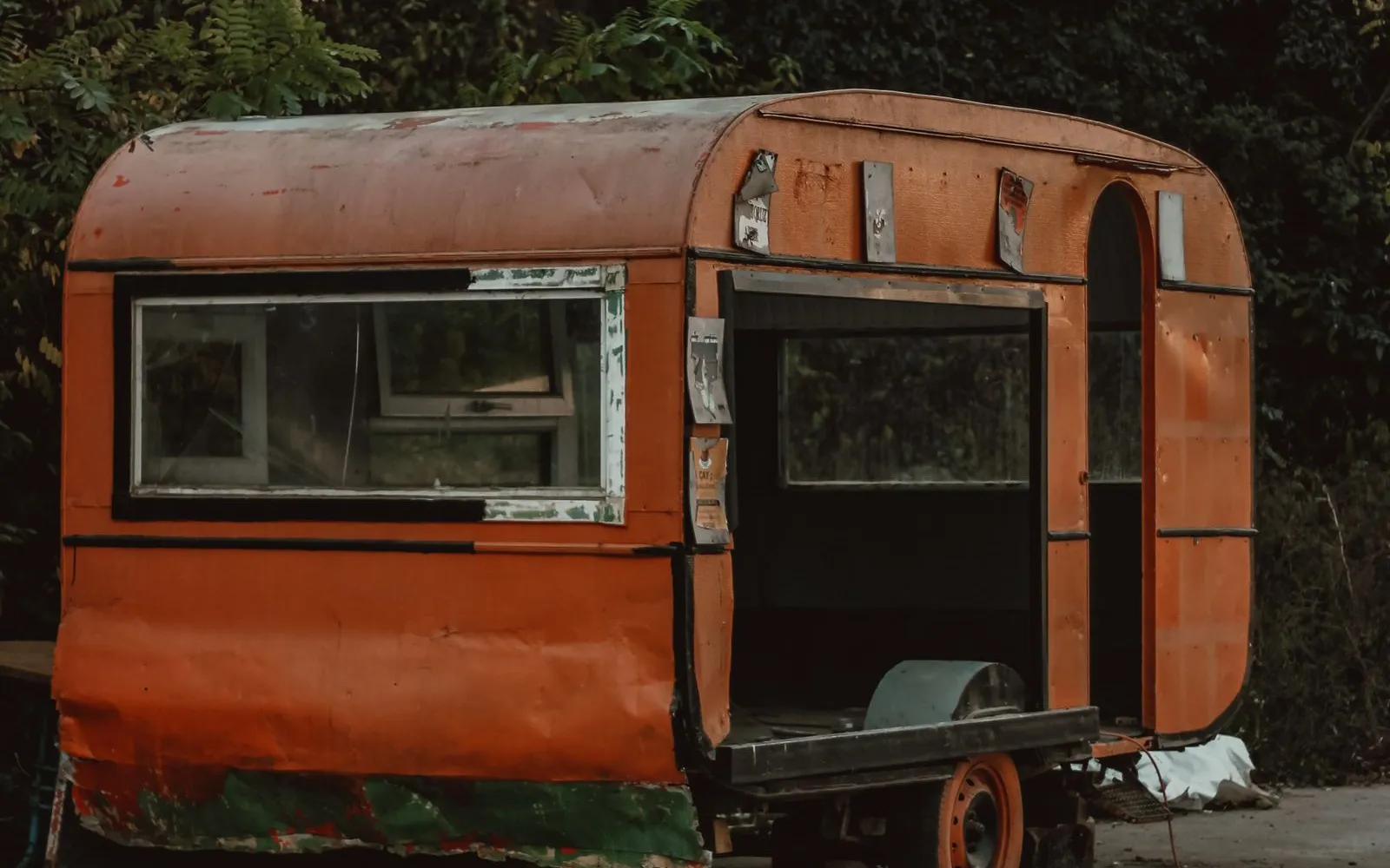 Weathered orange camping trailer with a missing door panel, parked at the edge of a road under trees