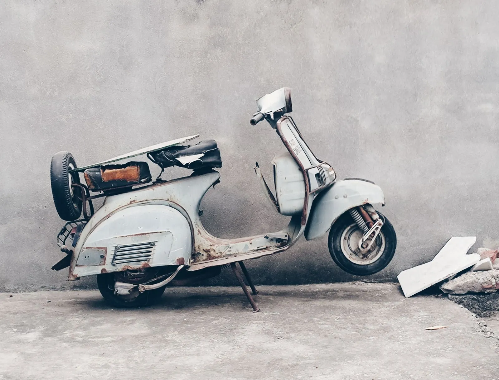 Rusted pale blue scooter with a spare wheel on the back, parked against a bare concrete wall